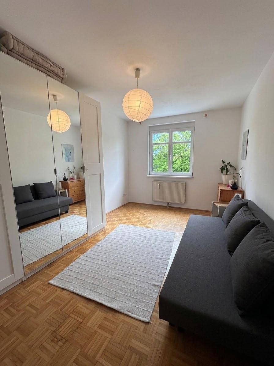 Geräumiges Zimmer mit Schlafsofa, großem Spiegelschrank und Parkettboden, hell durch das Fenster.