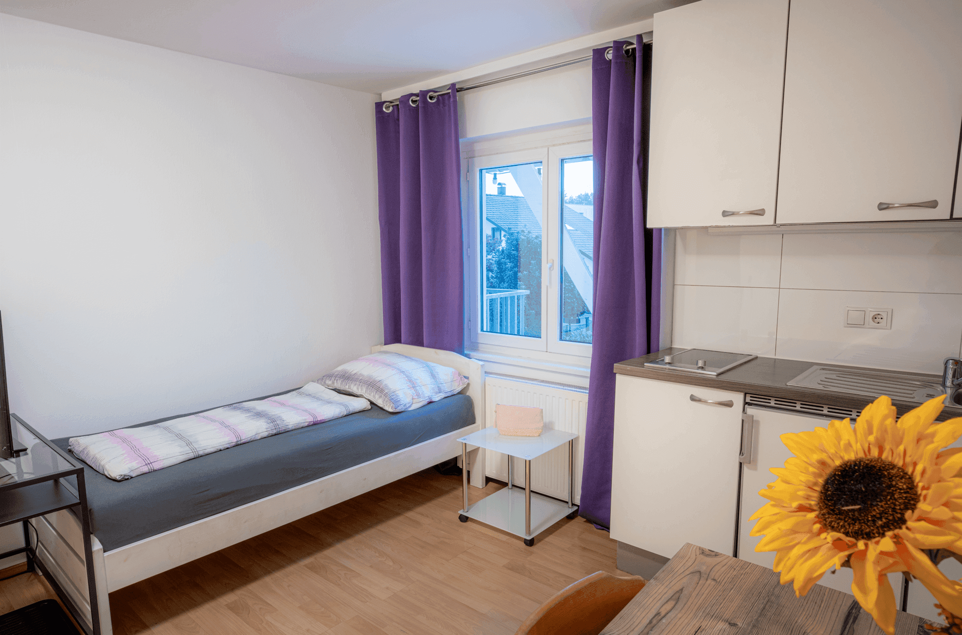 Kombinierter Wohn- und Schlafbereich mit Einzelbett, kleiner Küchenzeile und Fenster.