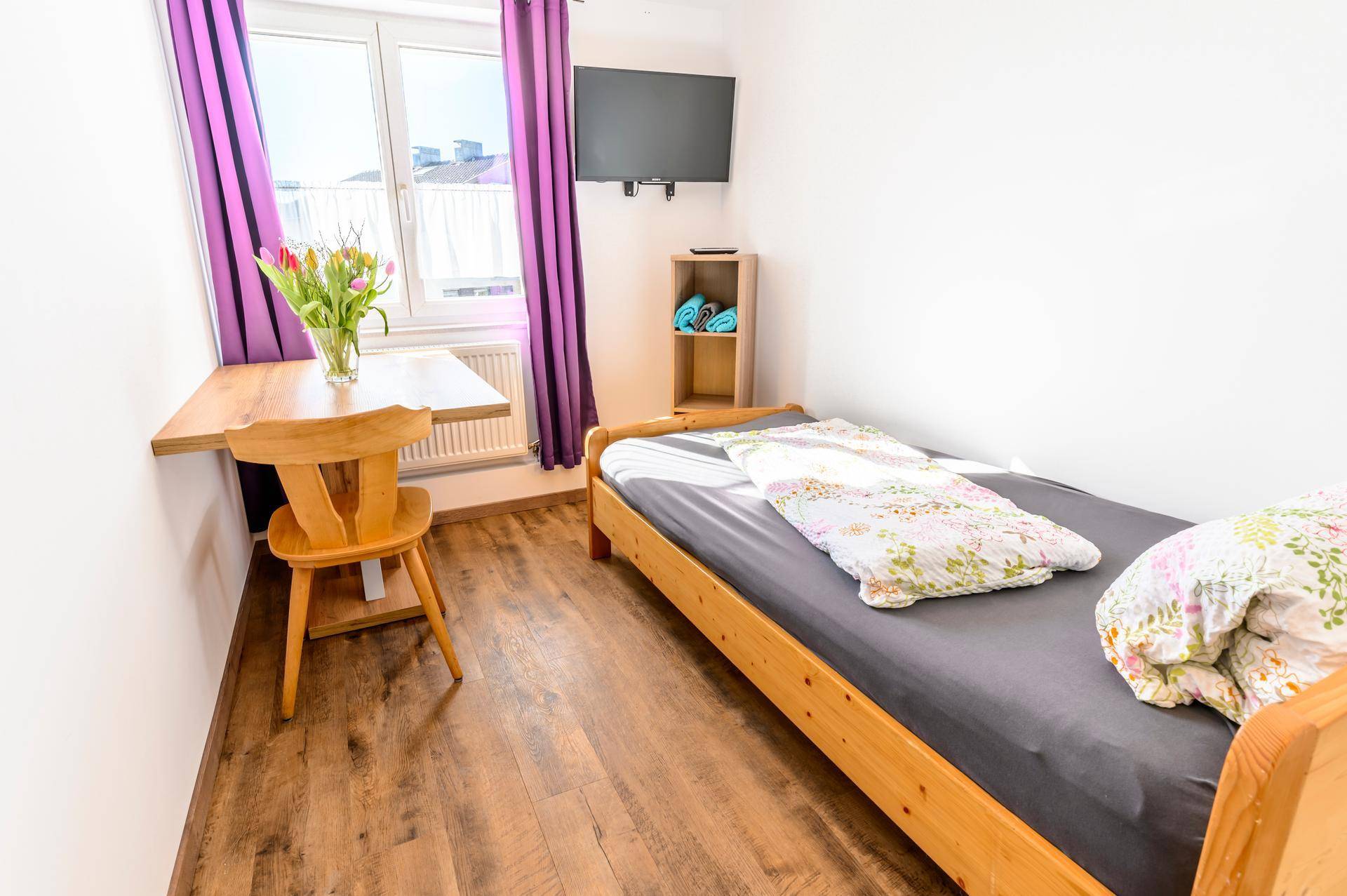 Kleines Schlafzimmer mit Einzelbett, Holztisch, Fernseher und Fenster mit lila Vorhängen.