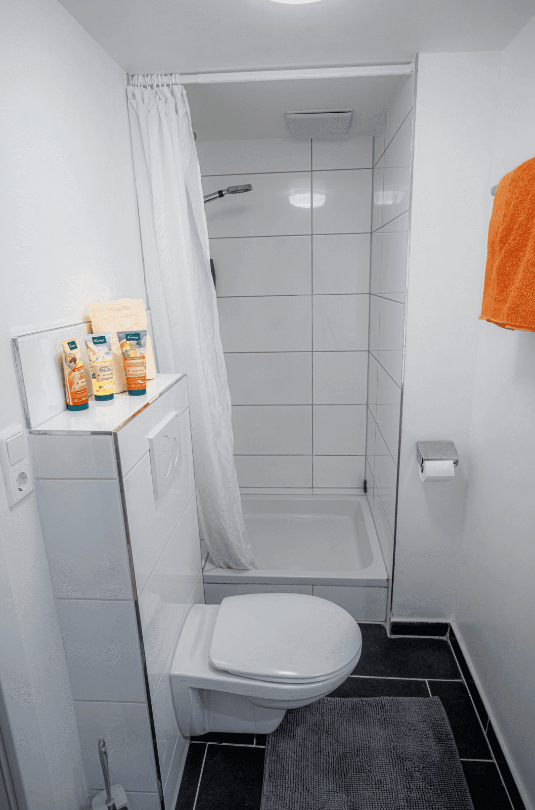 Badezimmer mit Toilette und Duschbereich, ausgestattet mit weißen Wandfliesen und dunklem Boden.