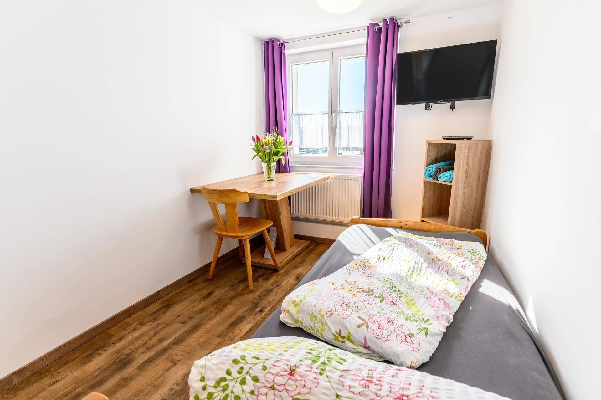 Gemütliches Schlafzimmer mit Einzelbett, Holztisch, Fernseher und Fenster mit lila Vorhängen.
