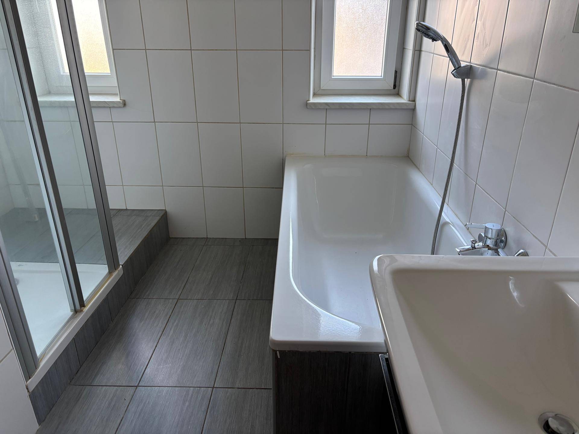 Helles Badezimmer mit Badewanne, Duschbereich und Fenster, ausgestattet mit weißen Fliesen.