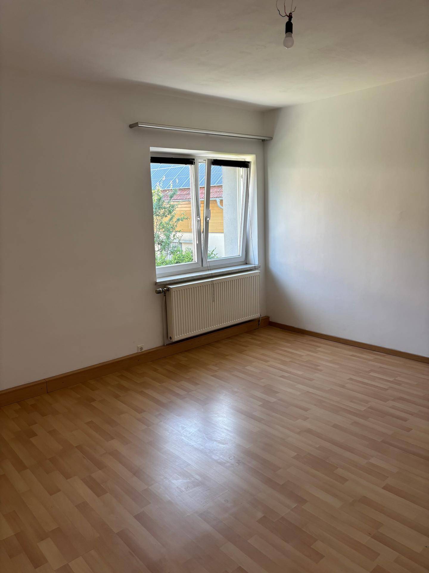 Leeres Zimmer mit Laminatboden und großem Fenster, das viel Tageslicht hereinlässt.