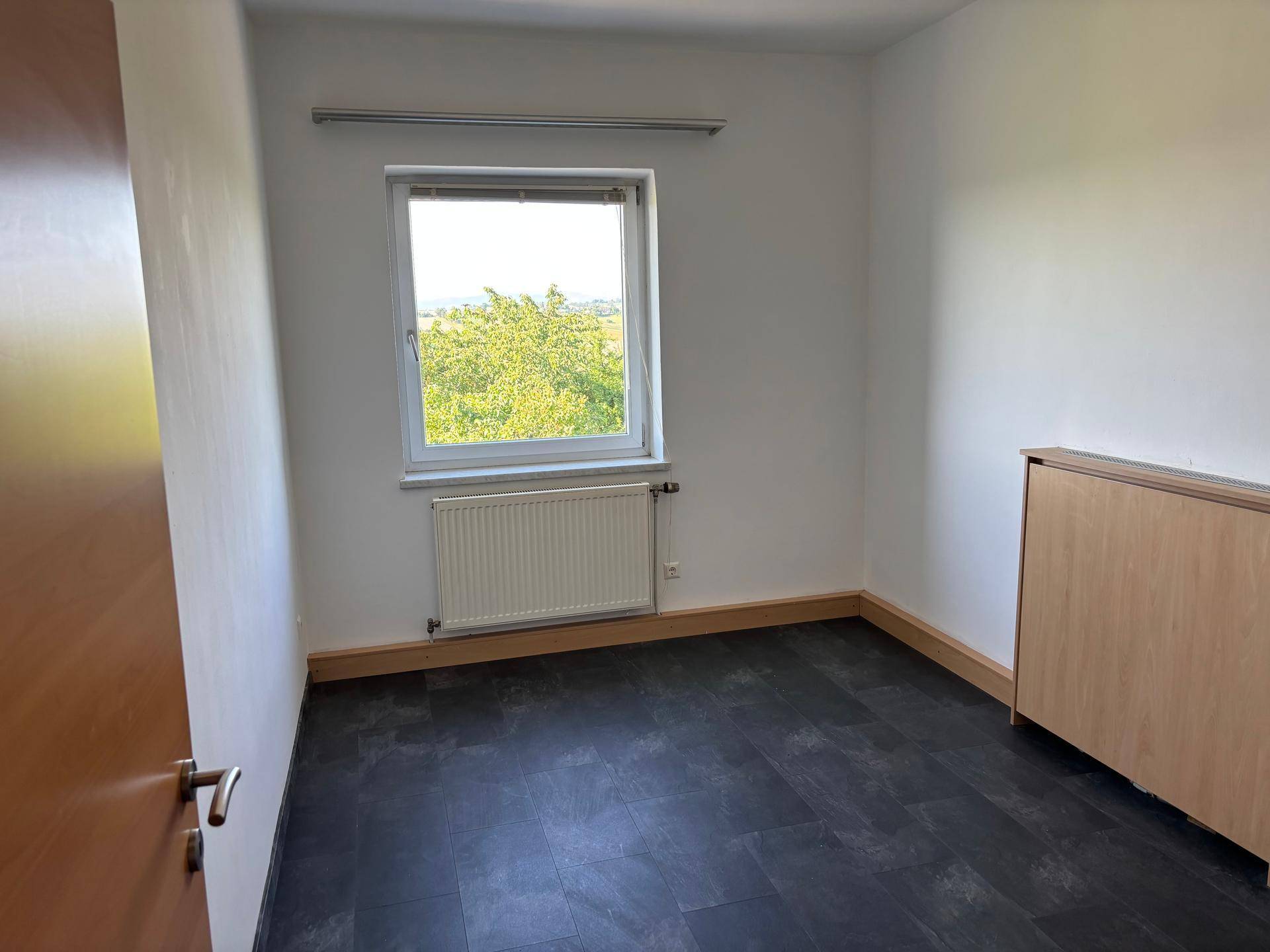 Helles Zimmer mit dunklem Vinylboden und Fenster mit Blick ins Grüne.