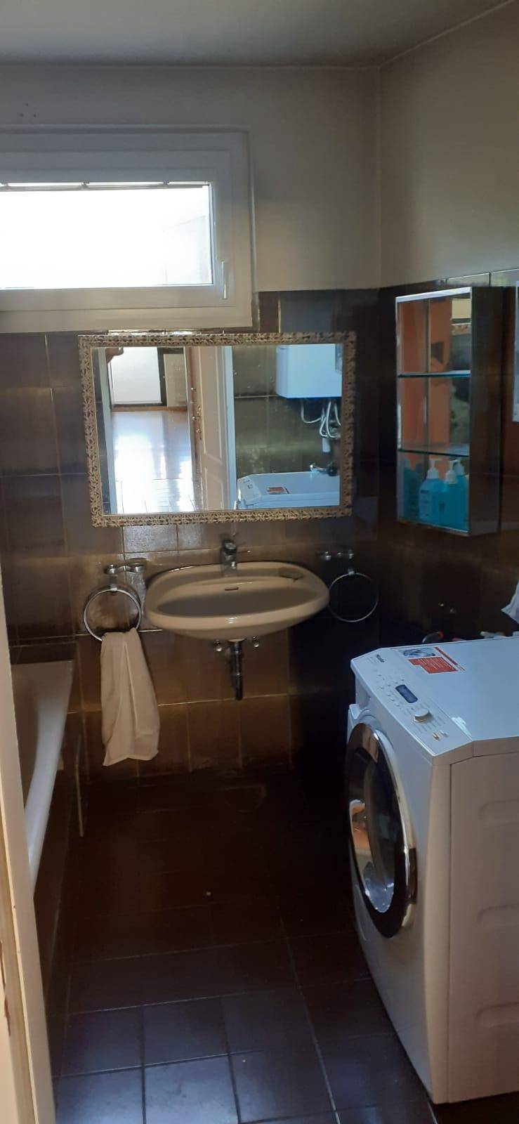 Badezimmer mit Waschbecken, Spiegel, Fenster und einer Waschmaschine, geflieste Wände und Boden.