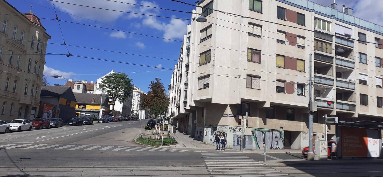 Stadtansicht mit Gebäuden, Straße und Fußgängerüberweg an einem sonnigen Tag.