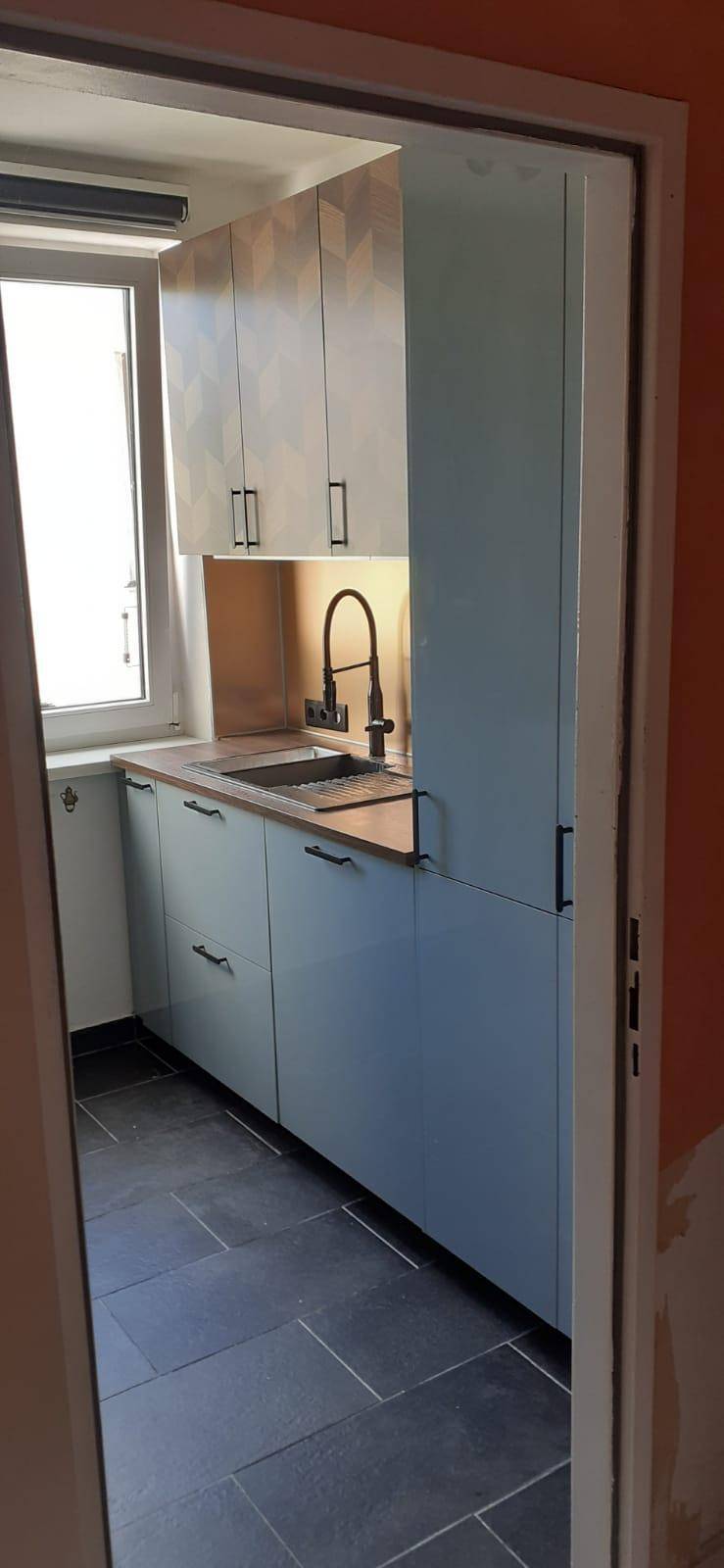 Kompakte Küche mit Fliesenboden, hellblauen Unterschränken und Fenster für Tageslicht.