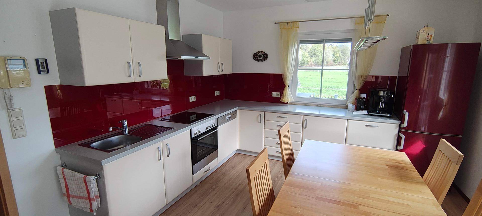 Helle Küche mit weißen Schränken, roter Rückwand, Holztisch und Blick ins Grüne.