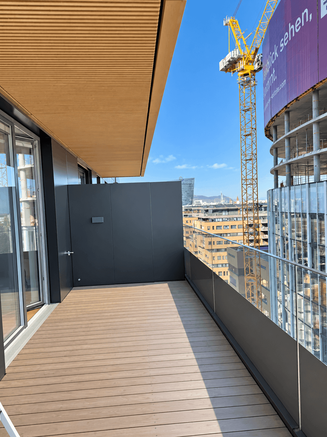 Langer Balkon mit Holzdielen und Blick auf eine Baustelle und die Stadtkulisse.