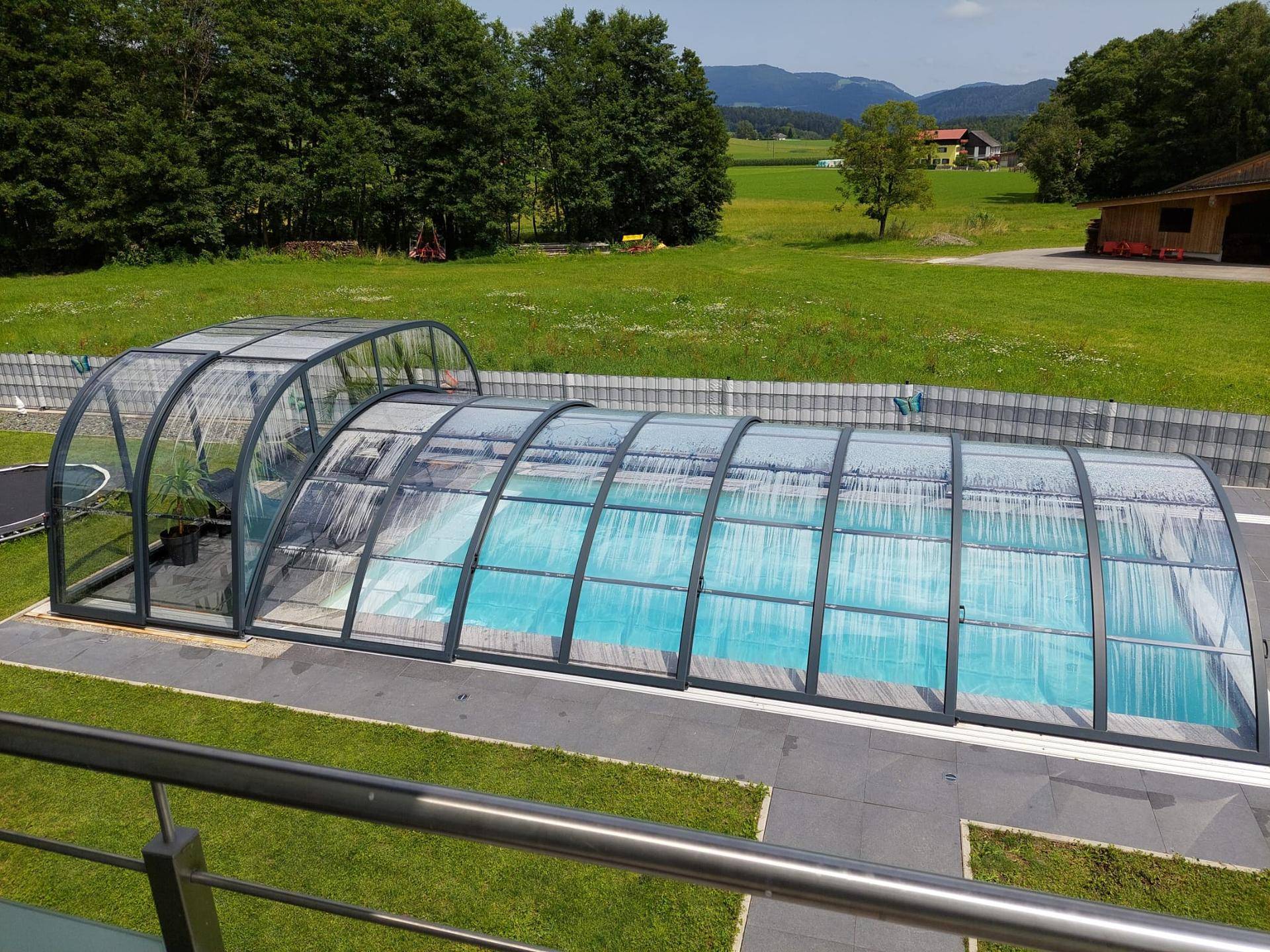 Großer, überdachter Swimmingpool im Garten, umgeben von einer gepflasterten Fläche und Bäumen.