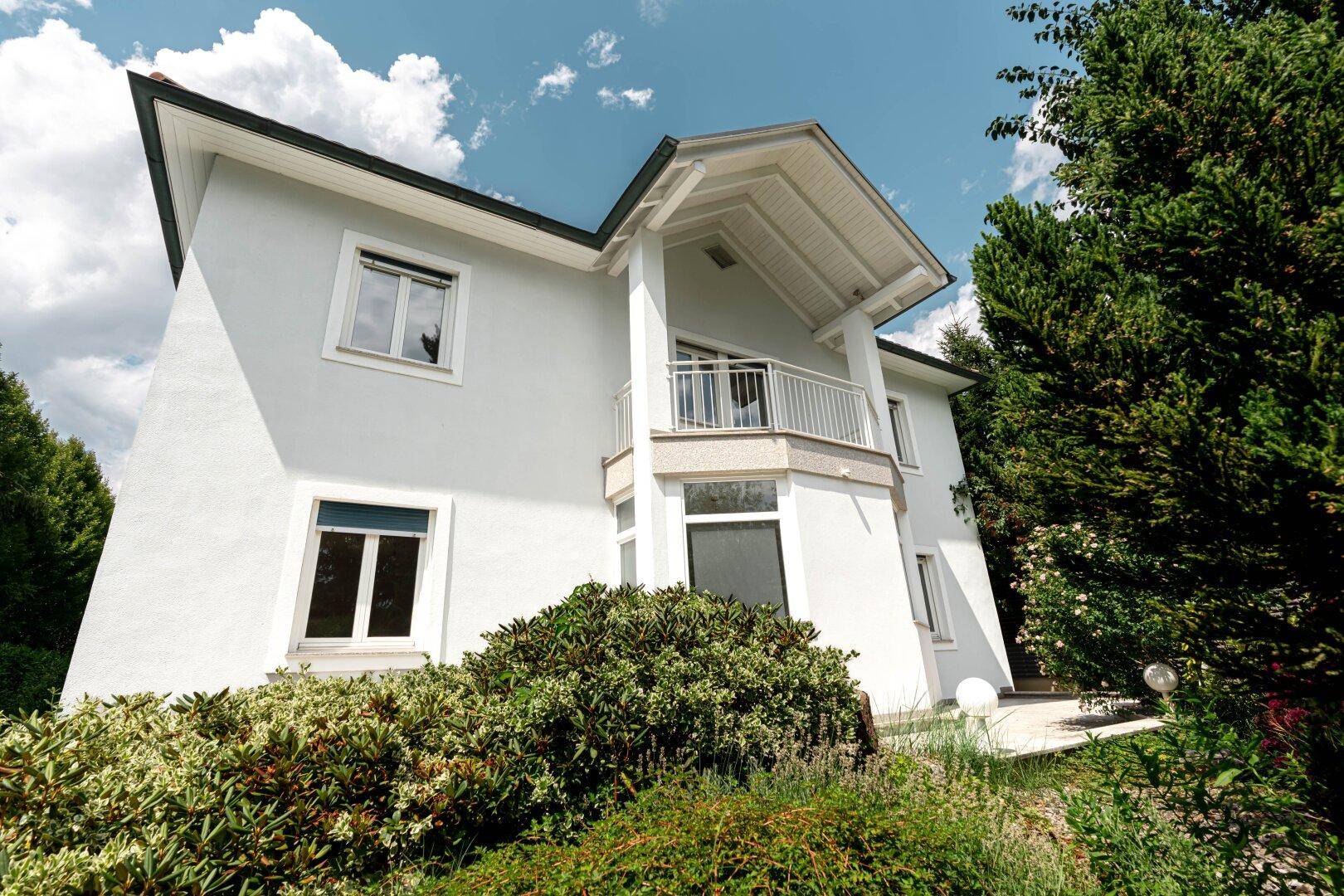 Moderne Hausfassade mit Balkon und großen Fenstern, umgeben von gepflegter Gartenlandschaft.