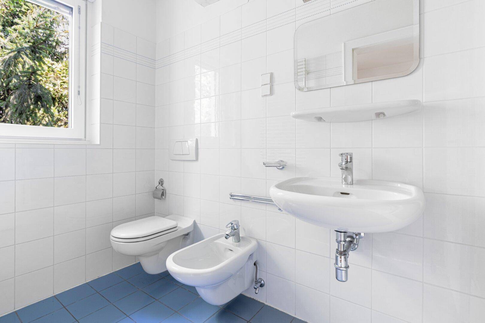Helles Badezimmer mit blauen Bodenfliesen, weiß gefliesten Wänden, WC, Bidet und Fenster.