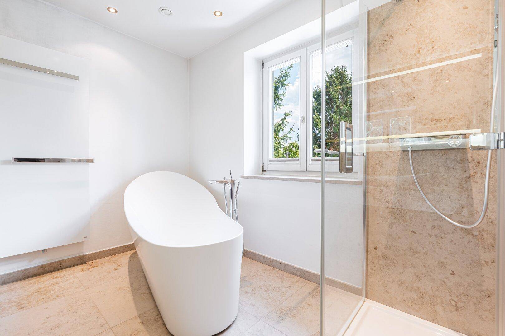 Elegantes Badezimmer mit freistehender Badewanne, großer Dusche und Fenster, das viel Tageslicht hereinlässt.