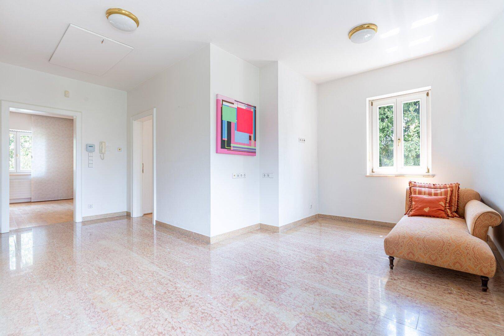 Helles Wohnzimmer mit Terrazzoboden, weiß gestrichenen Wänden und einem Fenster mit Blick ins Grüne.