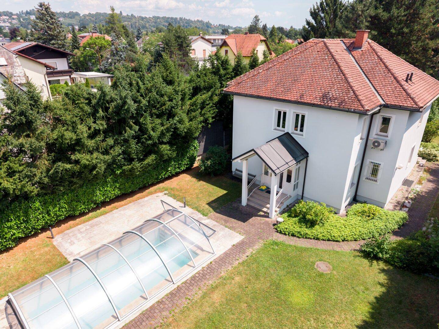 Luftaufnahme eines weißen Hauses mit rotem Ziegeldach, umgeben von einem grünen Garten und einem überdachten Pool.