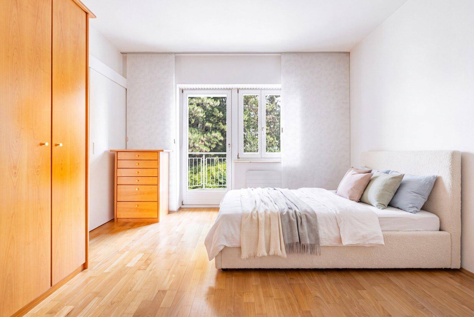 Helles Schlafzimmer mit Holzboden, weißem Bett und Zugang zu einem französischen Balkon mit Blick ins Grüne.