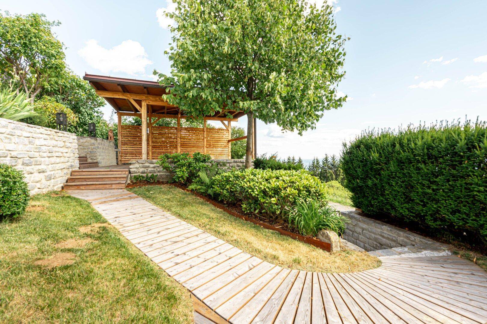 Überdachter Sitzbereich im Garten mit Holzweg und Steinmauern, ideal zum Entspannen im Freien.
