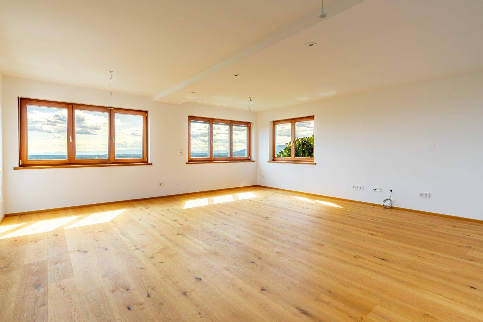 Geräumiges, leeres Wohnzimmer mit hellem Holzboden und großen Fenstern, die viel Licht hereinlassen.