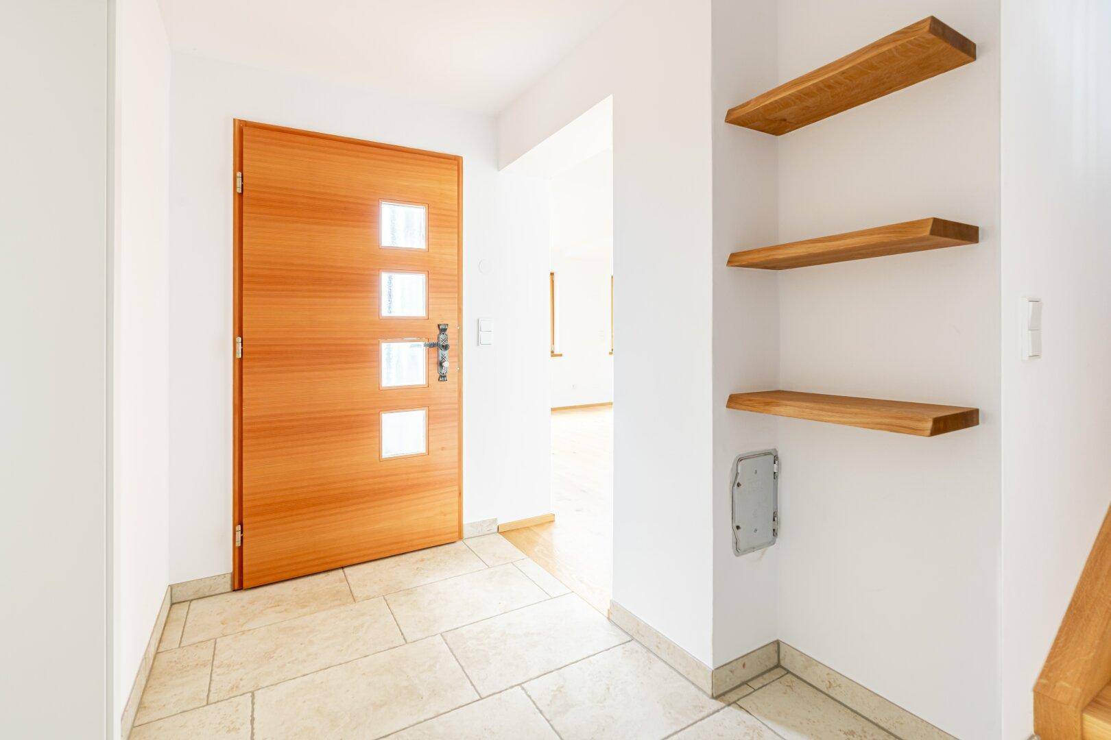 Heller Eingangsbereich mit Fliesenboden, Holztür und schwebenden Holzregalen an der Wand.