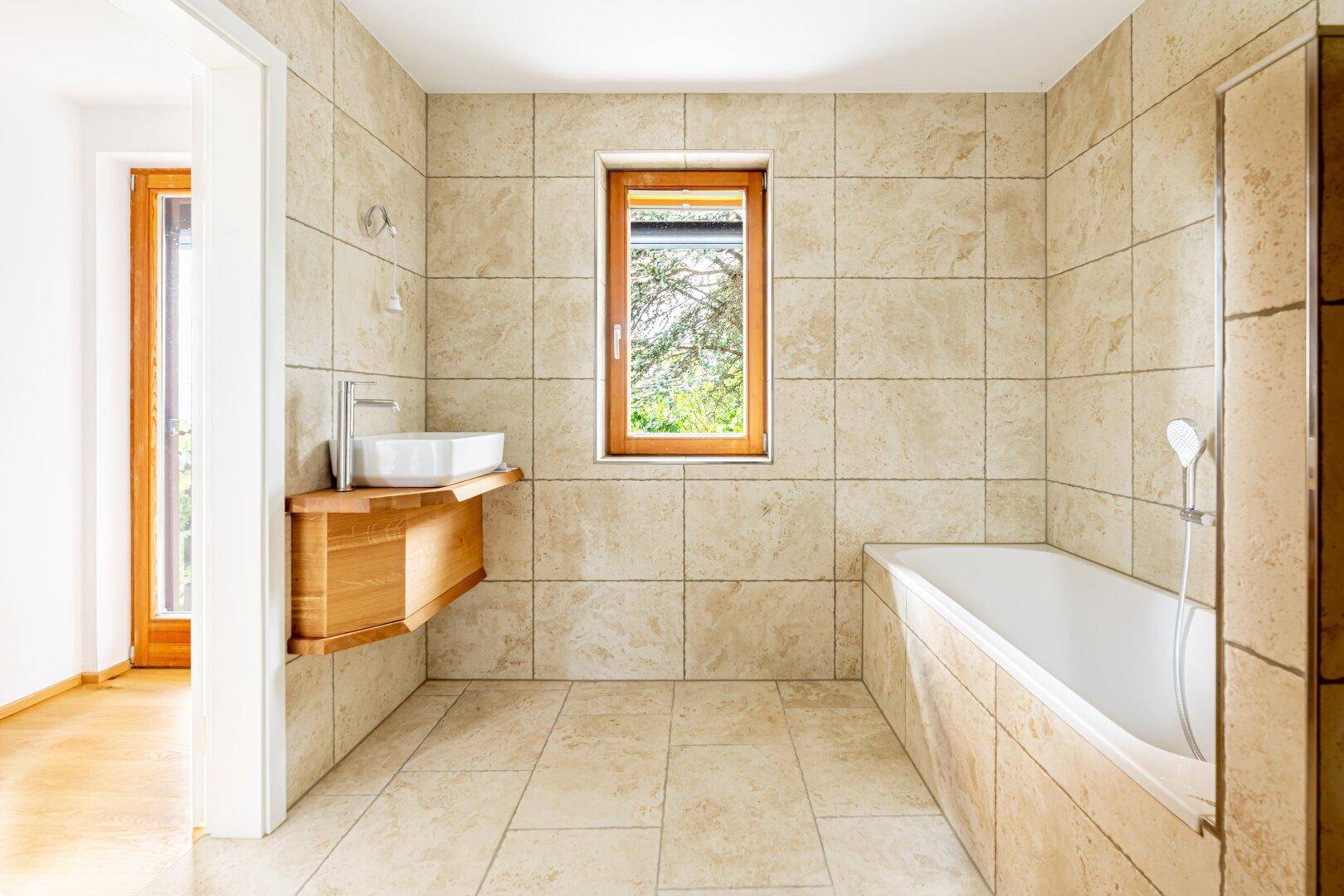 Helles Badezimmer mit beigefarbenen Fliesen, Badewanne, Holzwaschtisch und Fenster mit Ausblick.