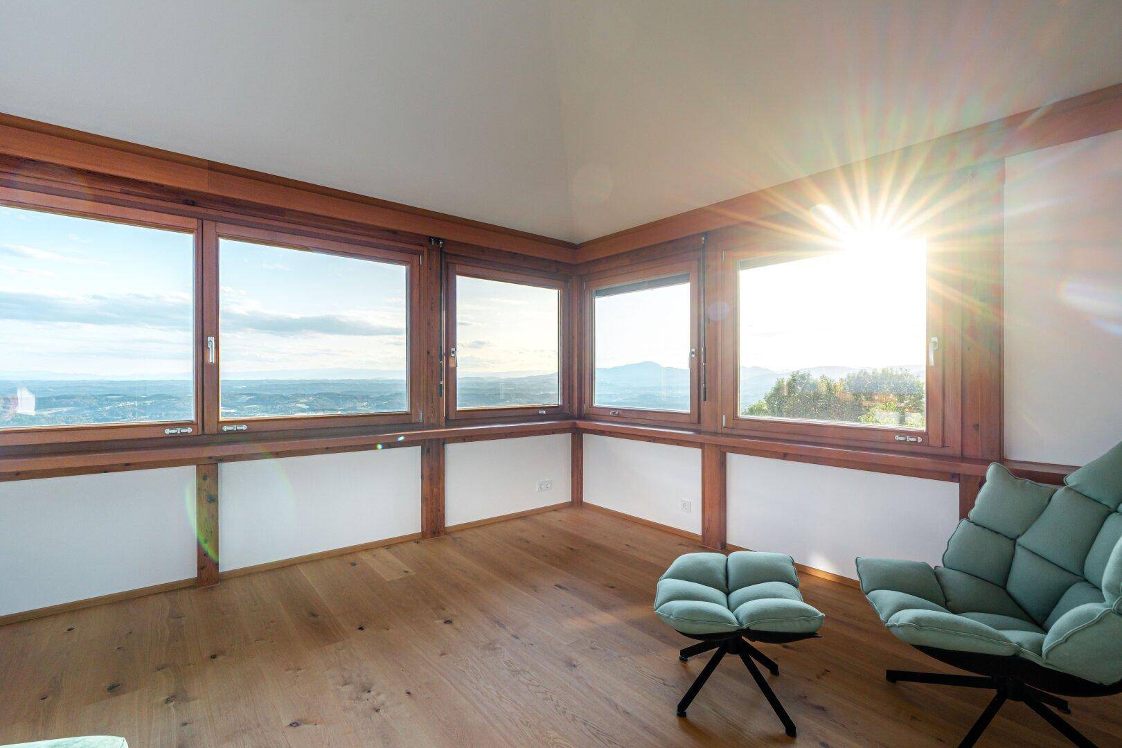 Lichtdurchfluteter Wohnbereich mit vielen Fenstern, Holzboden und Panoramablick über die Landschaft.