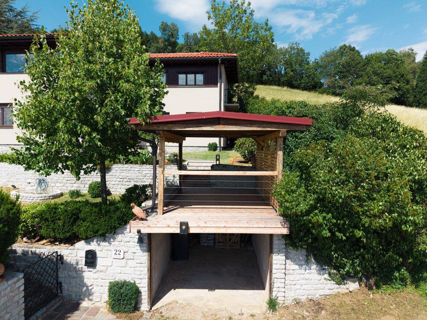 Hausfassade mit Holzterrasse über einer Garageneinfahrt, umgeben von grünen Büschen und Bäumen.