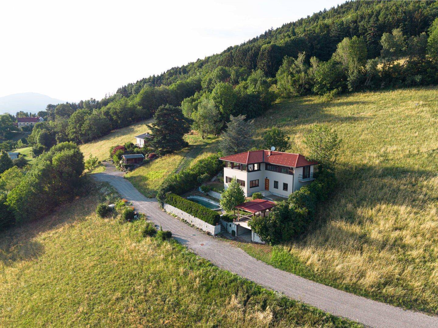 Luftaufnahme eines Hauses auf einem grünen Hügel mit Zufahrtsweg und umgebender Natur.