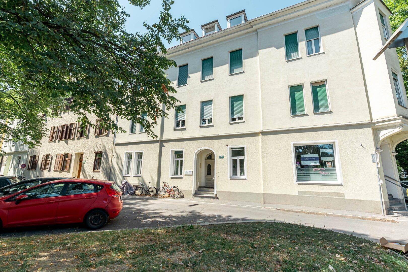 Mehrstöckiges Wohngebäude mit heller Fassade, grünen Fensterläden und geparkten Autos davor.