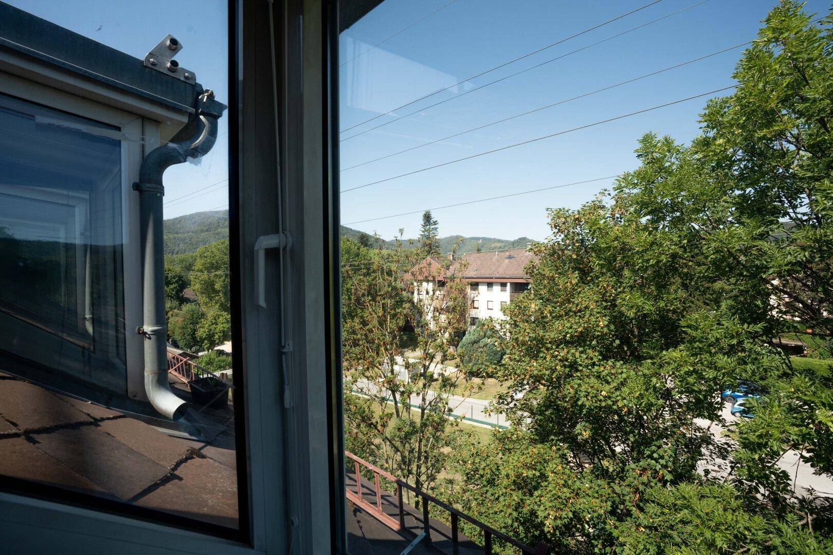 Ausblick aus dem Fenster auf grüne Bäume, umliegende Gebäude und sanfte Hügel in der Ferne.