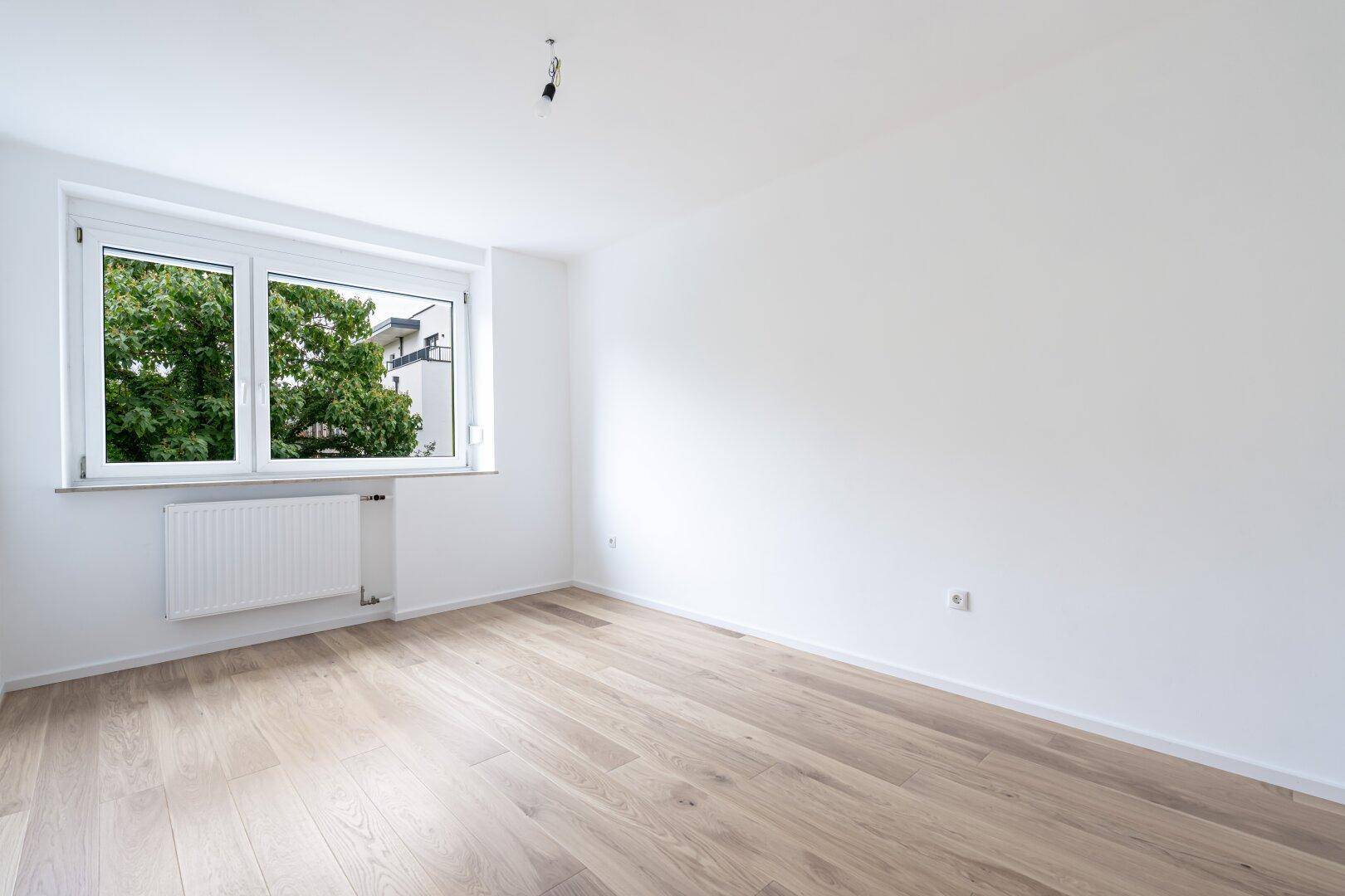 Helles, unmöbliertes Schlafzimmer mit Holzboden und großem Fenster mit Blick auf Bäume.