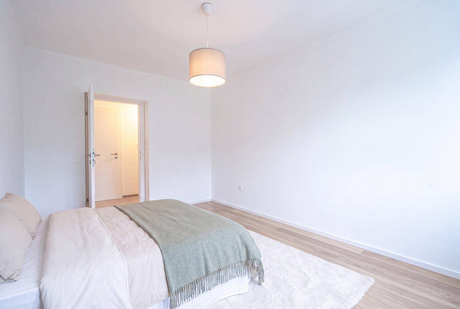Schlafzimmer mit hellem Holzboden und offener Tür, die den Blick in einen weiteren Raum freigibt.