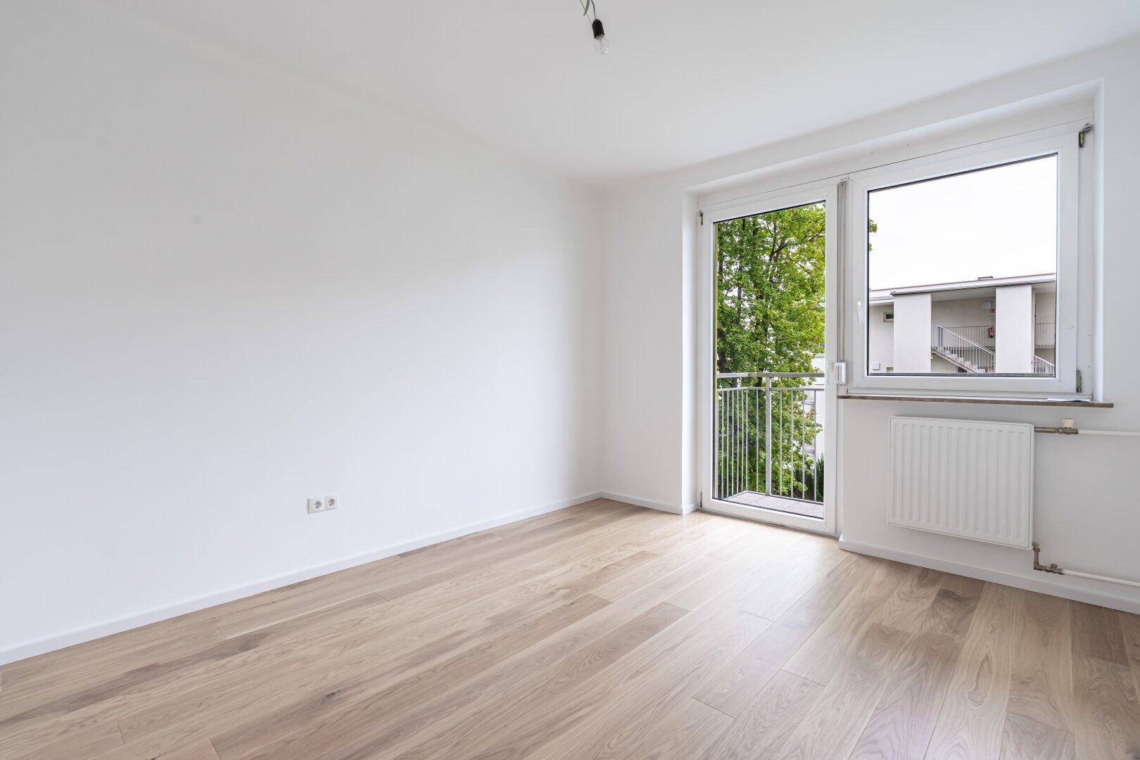 Unmöbliertes, helles Zimmer mit Holzboden und großem Fenster mit Zugang zum Balkon.