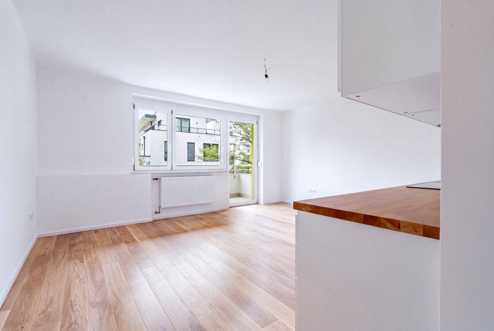 Heller, unmöblierter Wohn-Essbereich mit Küchenzeile, Holzboden und Zugang zum Balkon.