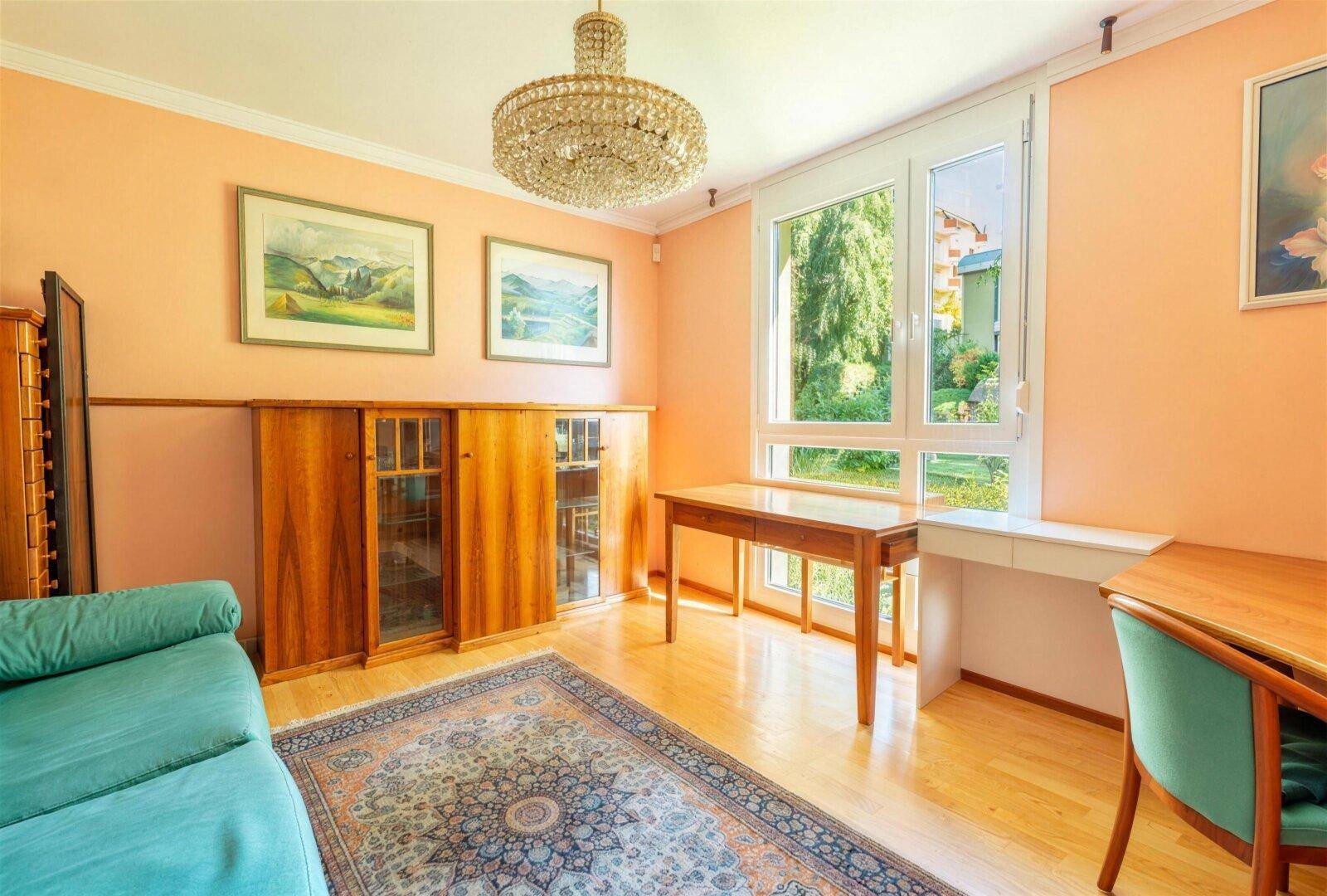 Arbeitszimmer mit Schreibtisch, Sofa, Kronleuchter und großem Fenster mit Grünblick.