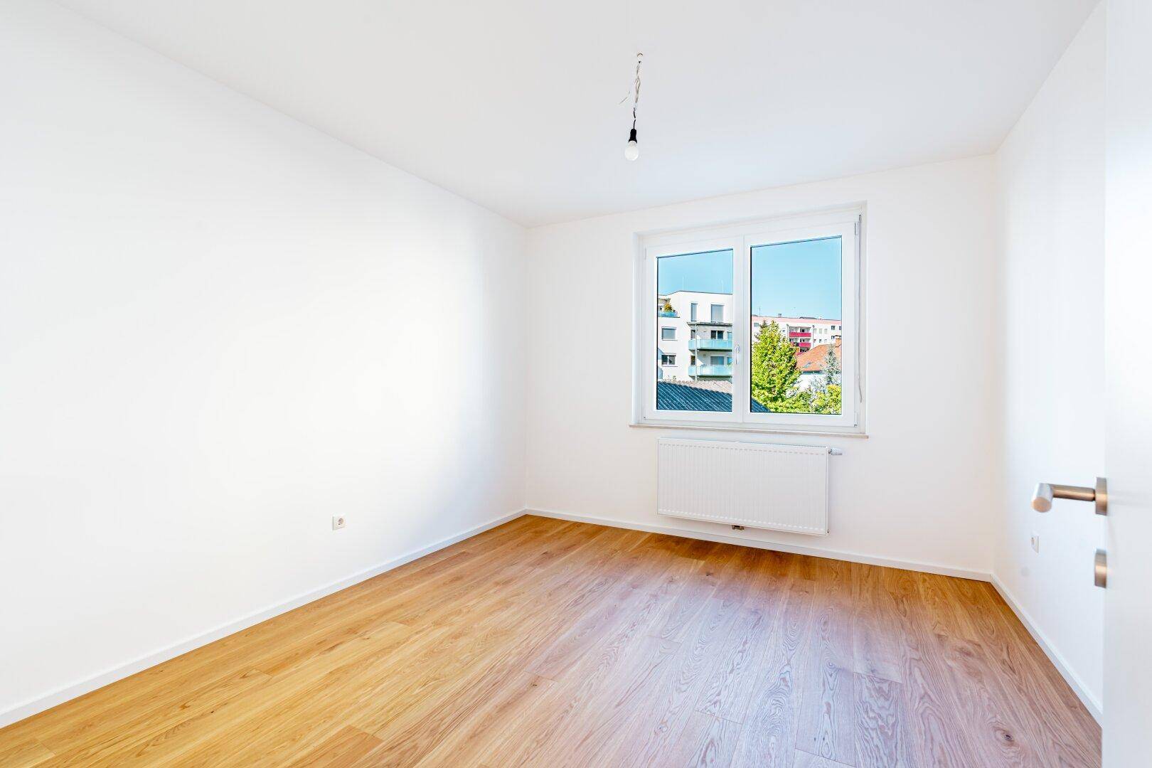 Helles, leeres Schlafzimmer mit Holzfußboden und einem Fenster, das den Blick auf andere Gebäude freigibt.