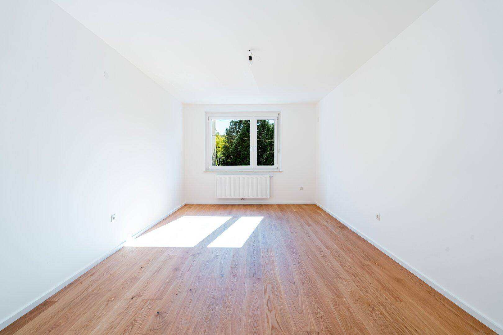 Leeres Schlafzimmer mit hellem Holzfußboden und einem Fenster, das viel Tageslicht in den Raum lässt.