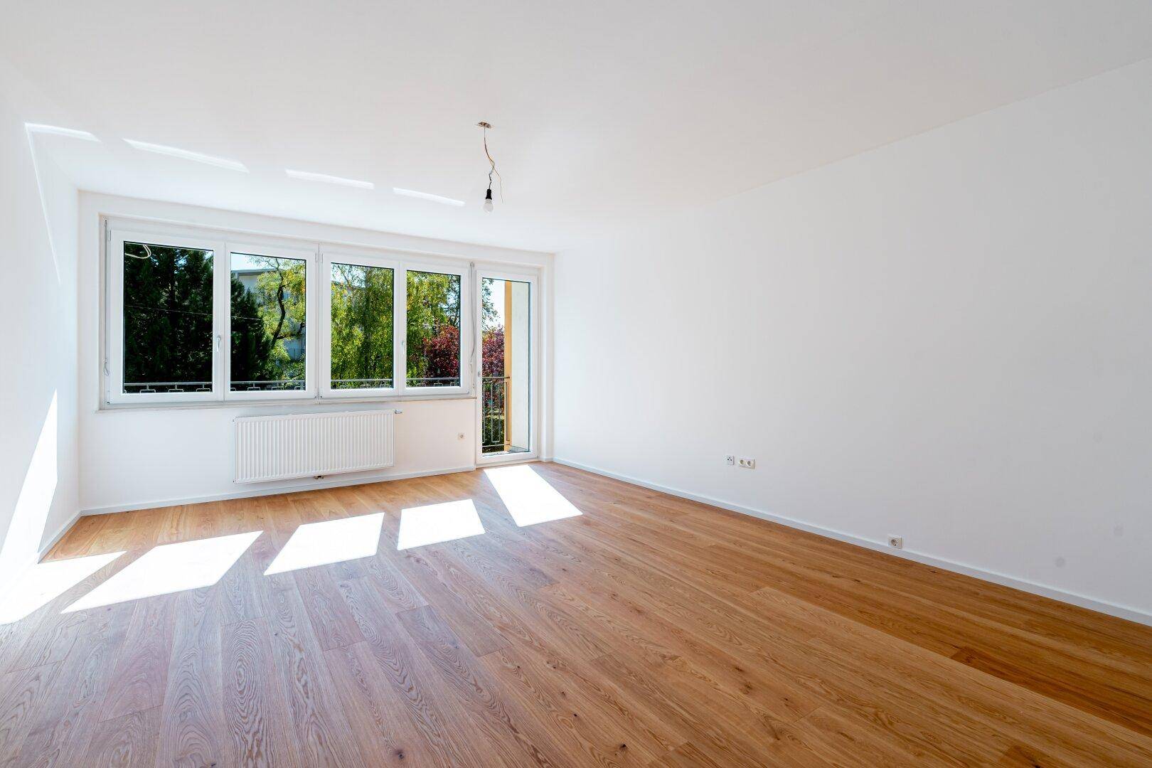 Geräumiger, heller Wohnbereich mit Holzfußboden und großen Fenstern, die einen Blick ins Grüne bieten.