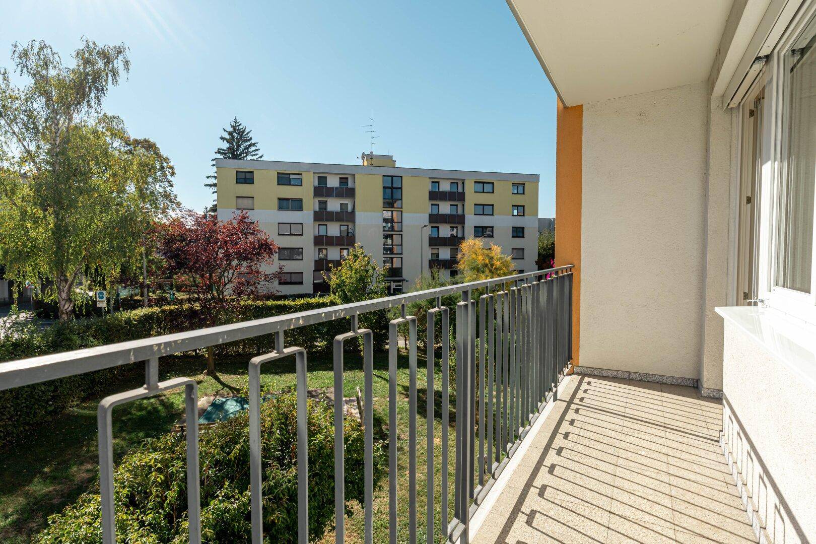 Balkon mit Blick auf einen grünen Innenhof und ein gegenüberliegendes Wohngebäude unter blauem Himmel.