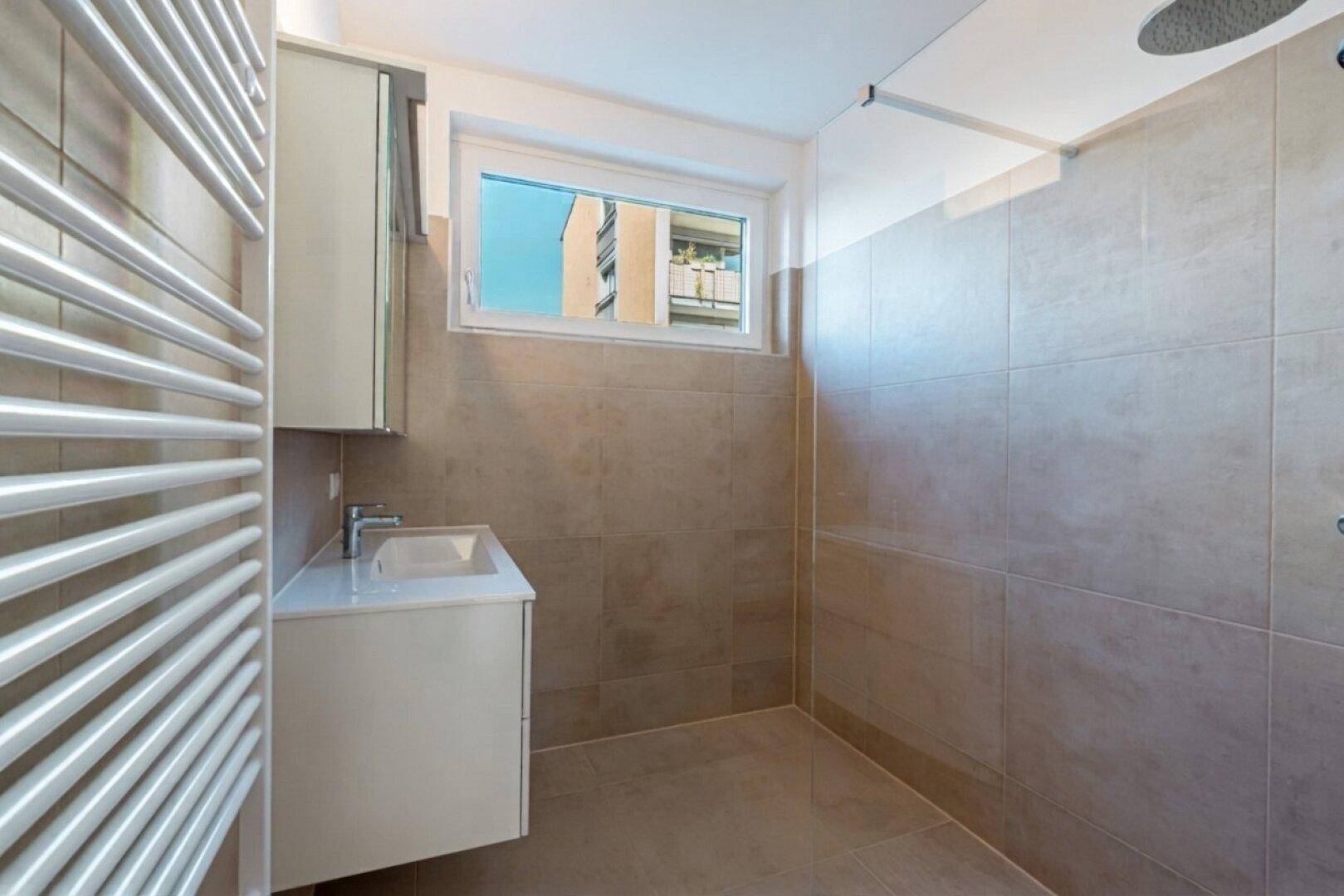 Modernes Badezimmer mit ebenerdiger Dusche, Einzelwaschbecken und beheiztem Handtuchhalter, hell gefliest.