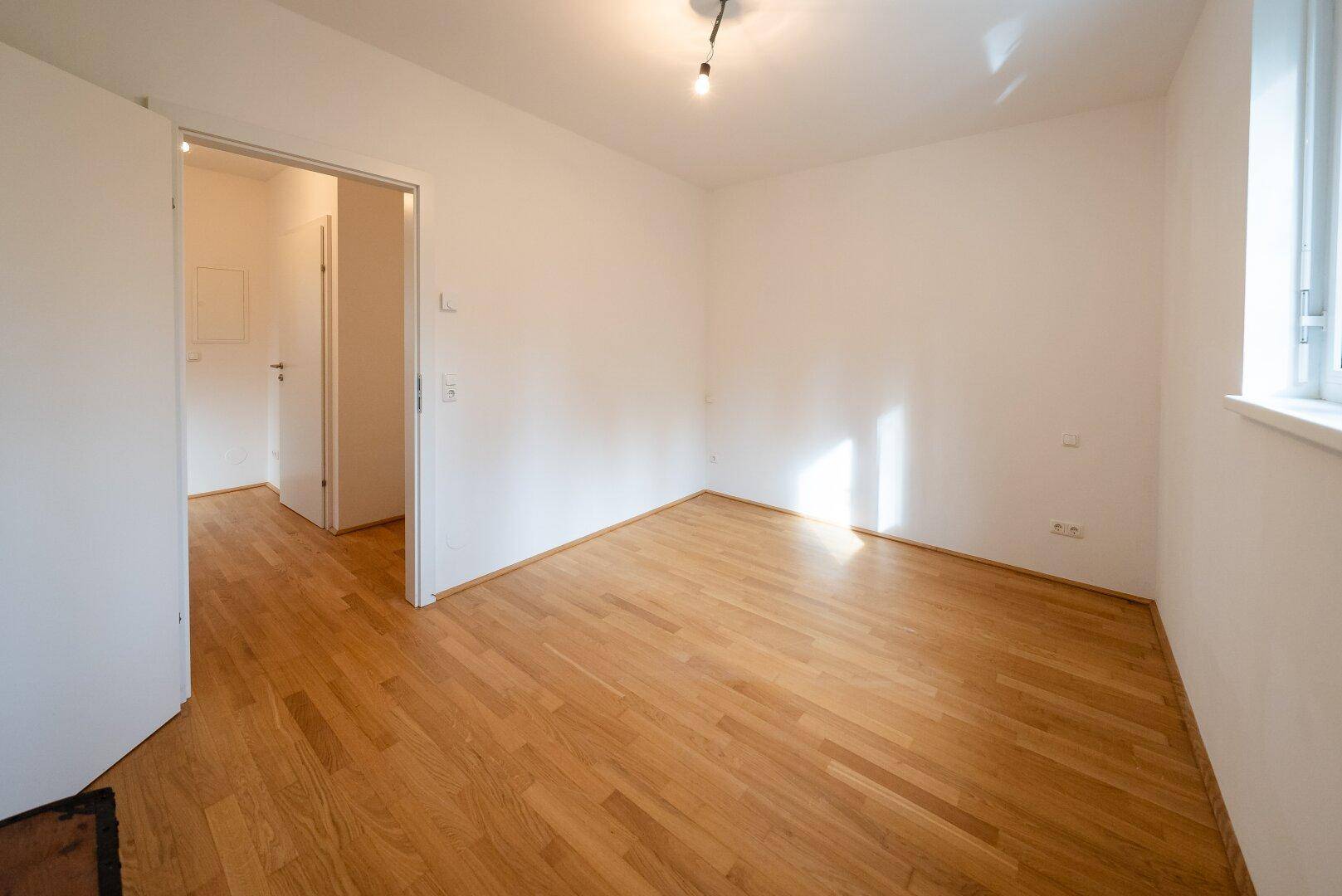 Leeres Zimmer mit Holzboden und Fenster, das viel Tageslicht hereinlässt.