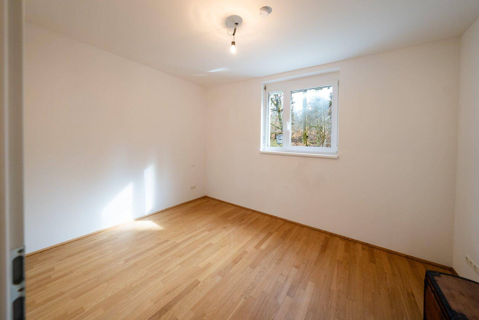 Helles Schlafzimmer mit Holzboden und Fenster, das einen Blick ins Grüne bietet.
