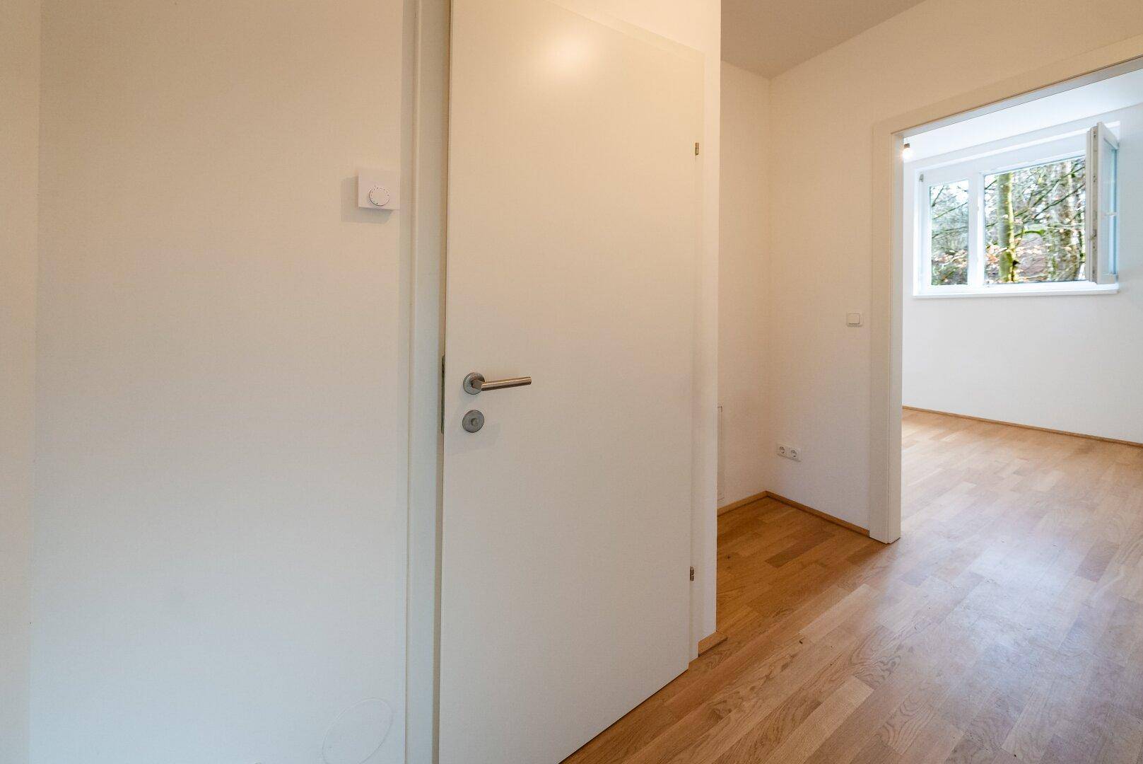 Weiß gestrichener Flur mit Holzboden und Zugang zu einem hellen Zimmer.
