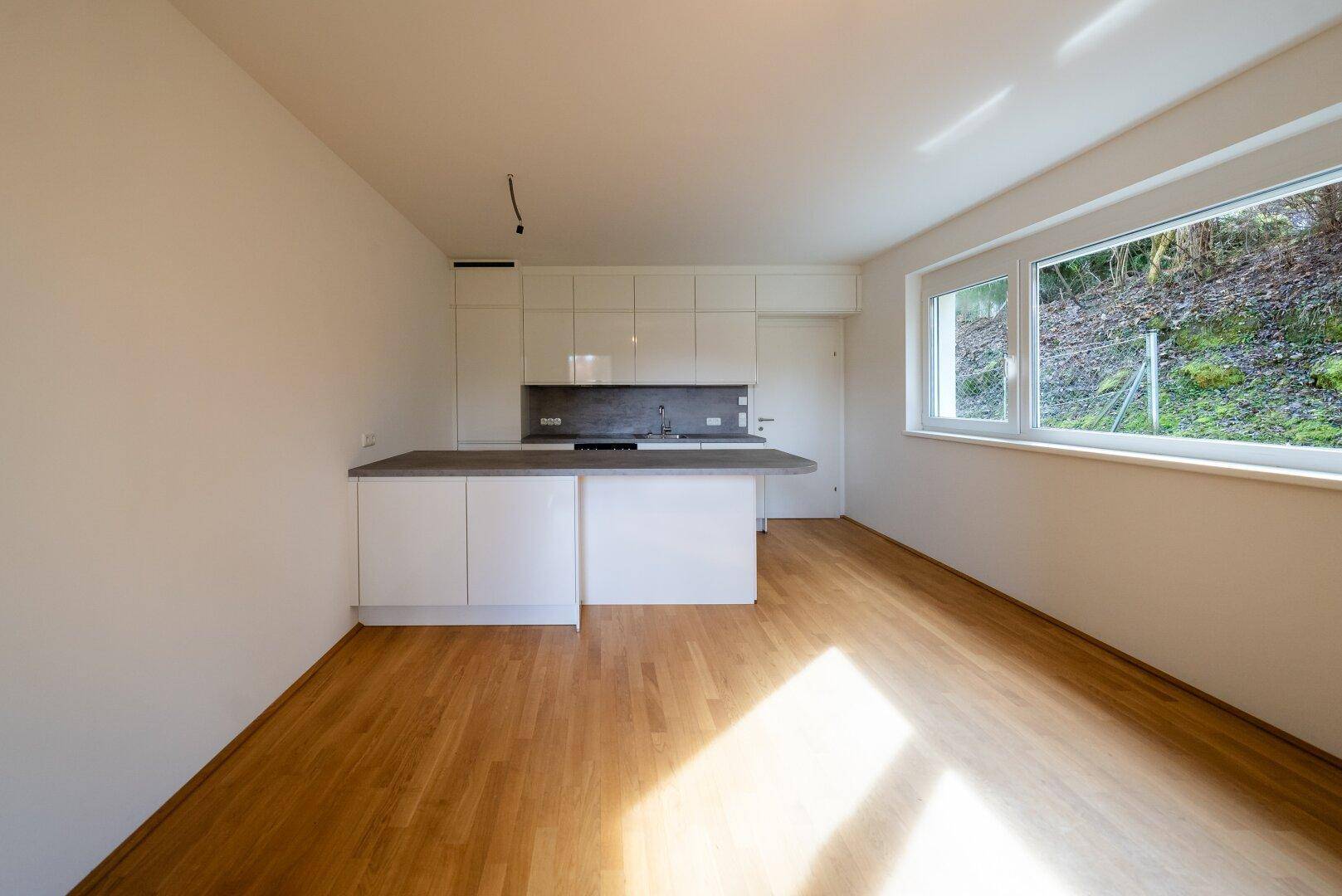 Geräumiger, leerer Wohn- und Küchenbereich mit Holzboden und großen Fenstern.
