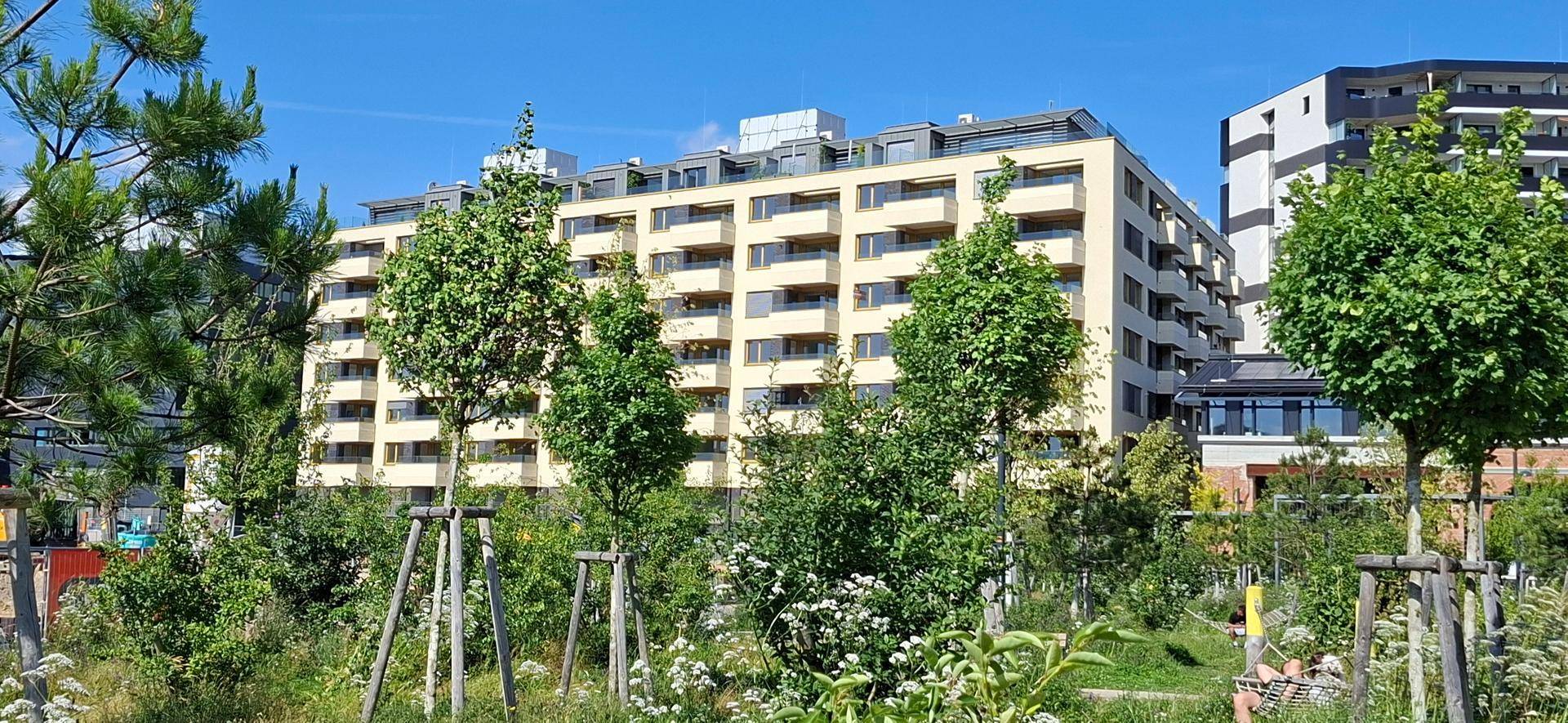 Modernes Wohngebäude mit vielen Balkonen, umgeben von grünen Bäumen unter blauem Himmel.