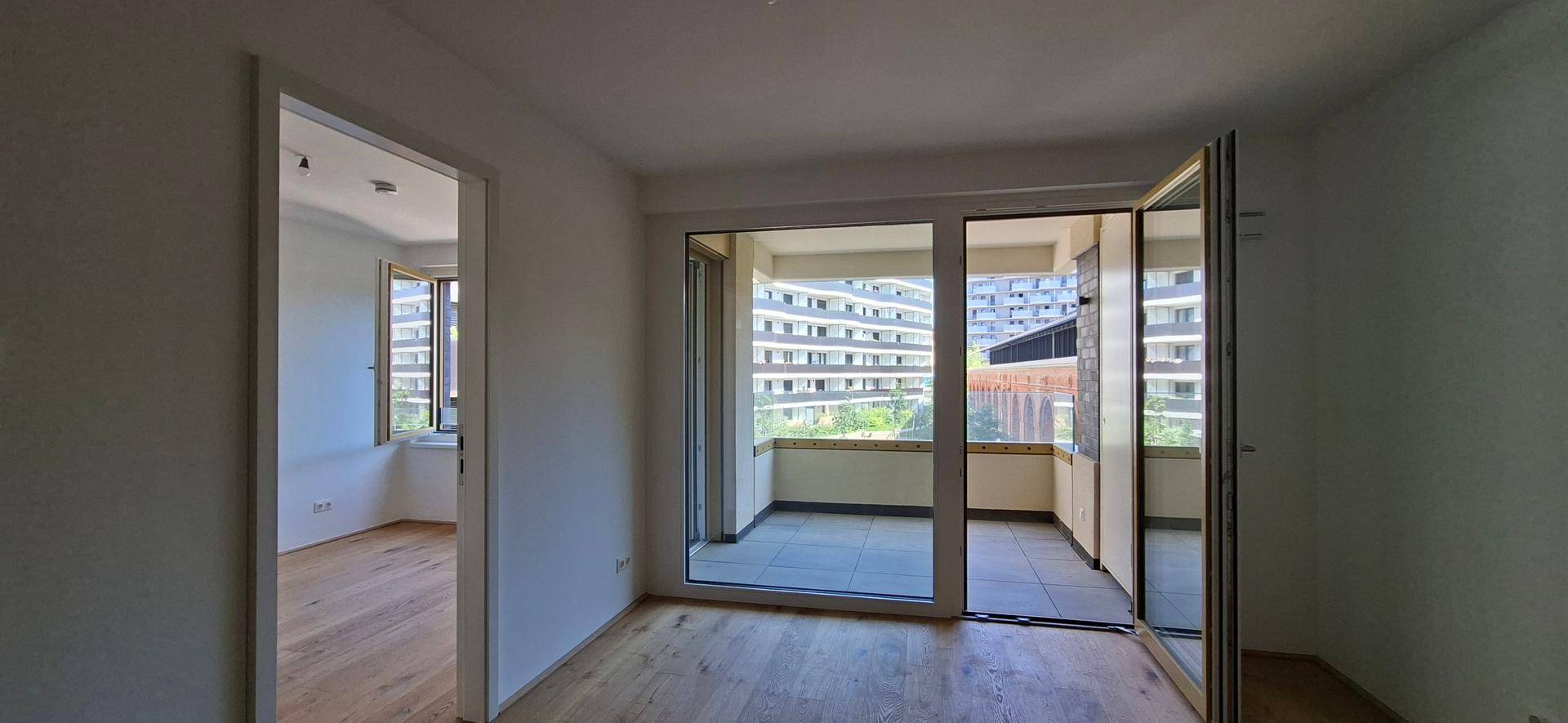 Heller Raum mit Holzboden und großen Fenstern, die Zugang zu einem Balkon bieten.