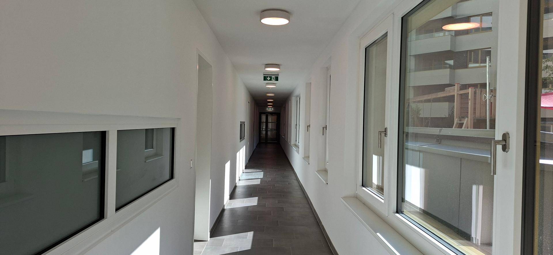 Langer, heller Flur mit Fliesenboden und Fenstern, die den Blick nach draußen freigeben.