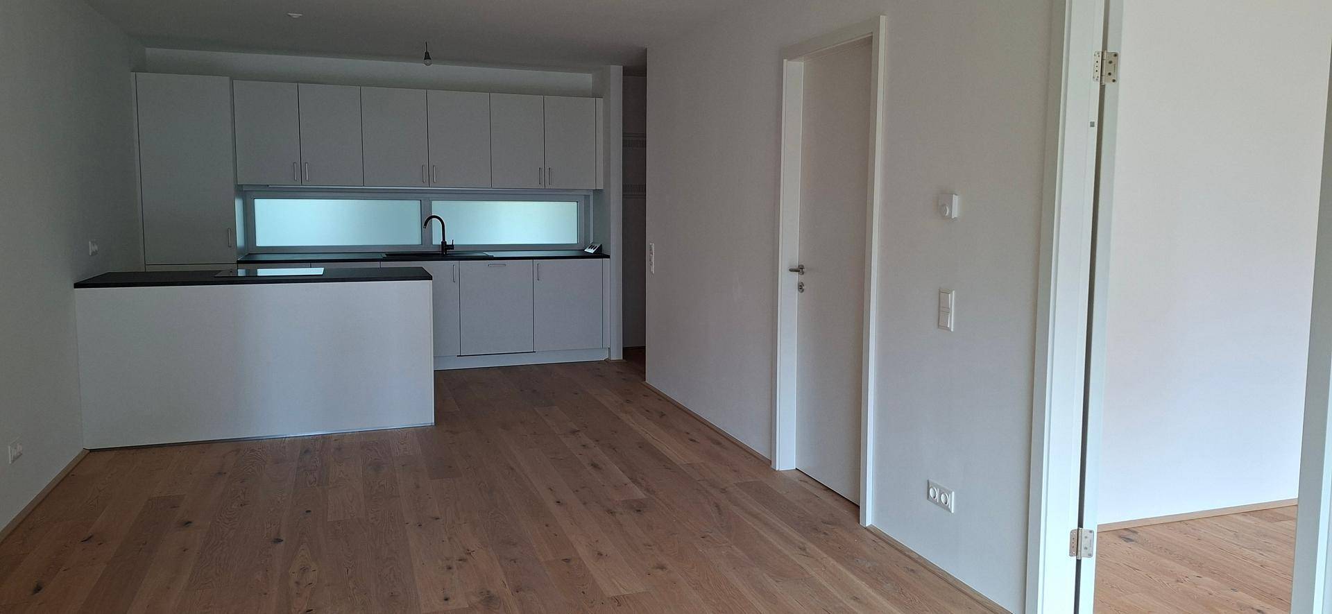 Offener Wohnbereich mit moderner weißer Küche, Kücheninsel und hellem Holzboden.