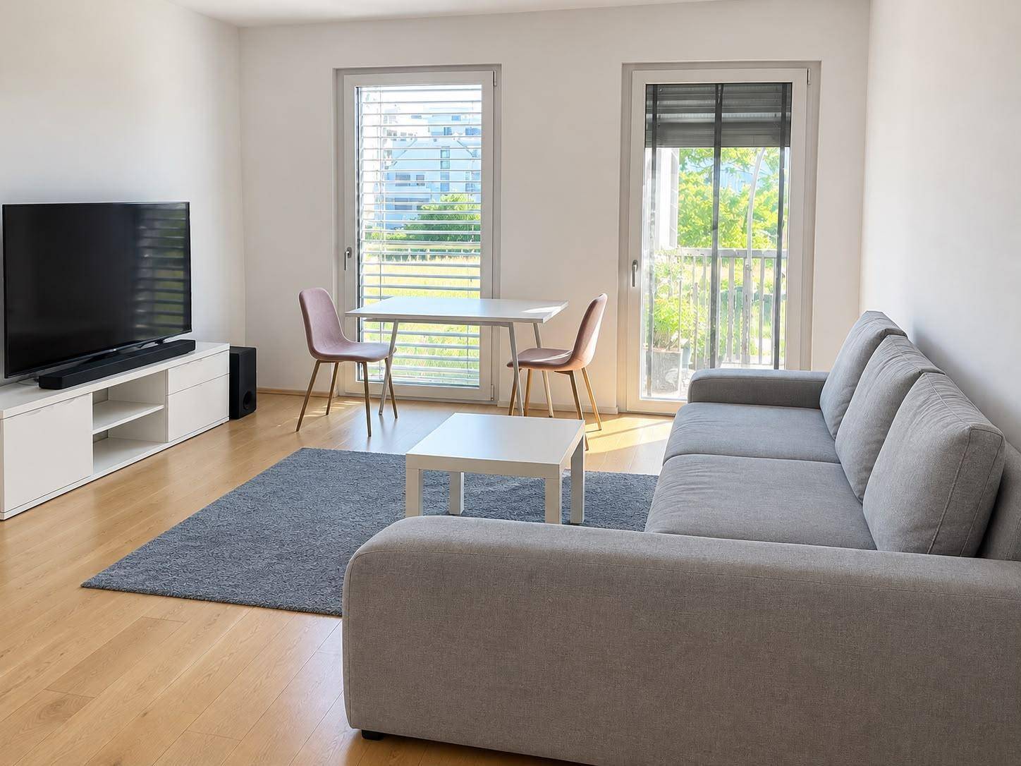 Heller Wohnbereich mit Sofa, Esstisch und Zugang zum Balkon durch große Fenster.