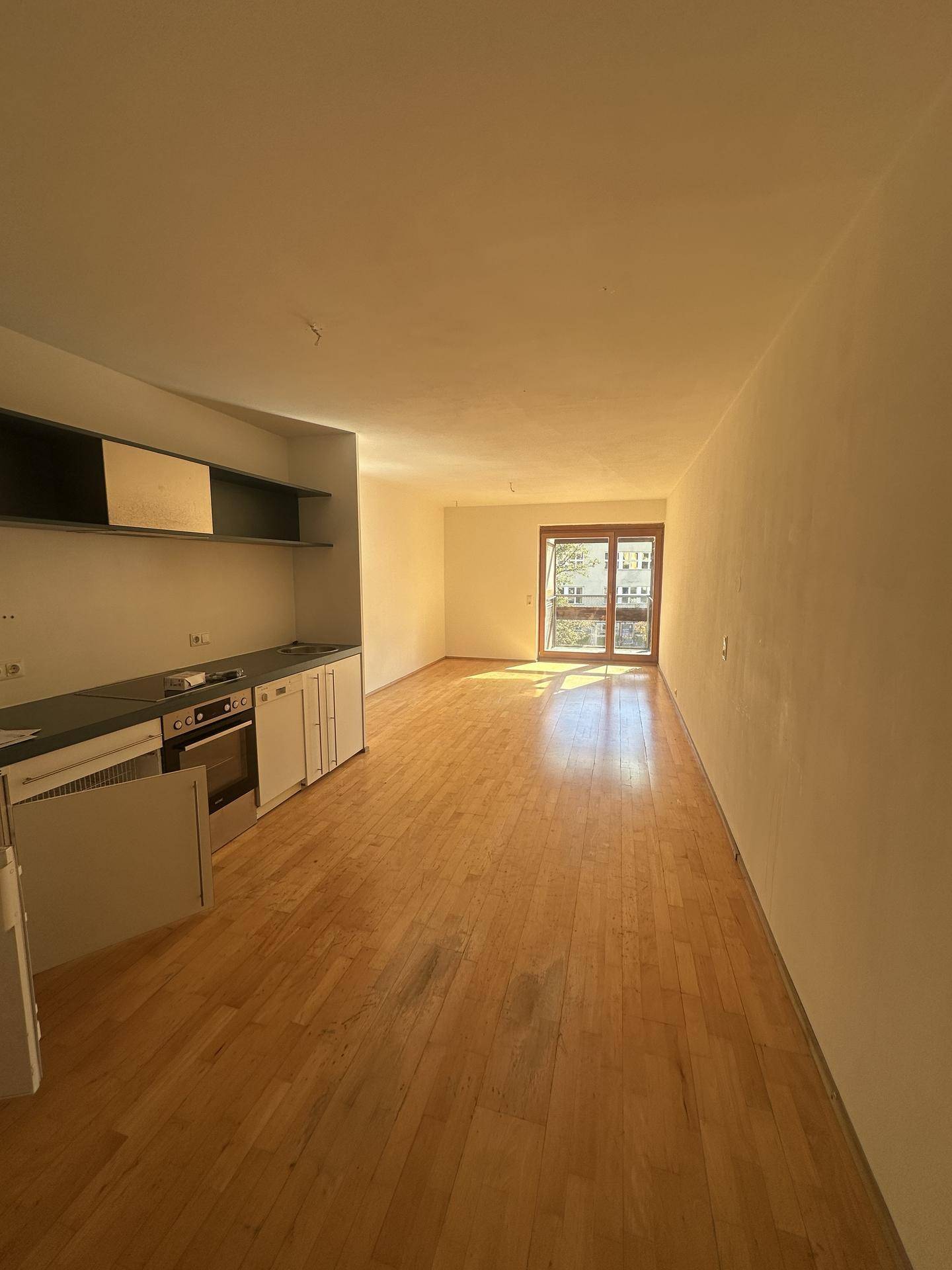 Offener Wohnbereich mit integrierter Küchenzeile, Holzboden und Zugang zu einem sonnigen Balkon.
