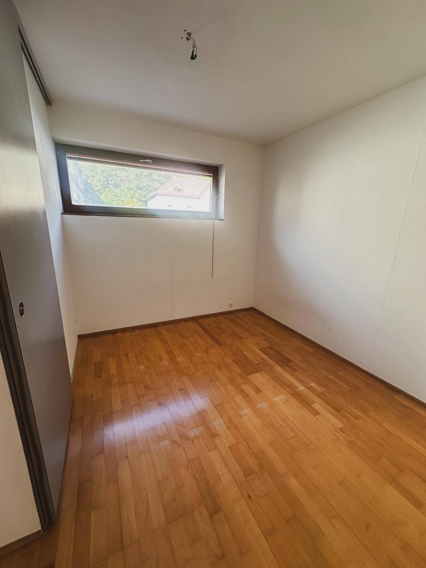 Ruhiges Zimmer mit Holzboden und einem Fenster, ideal als Schlaf- oder Arbeitszimmer nutzbar.
